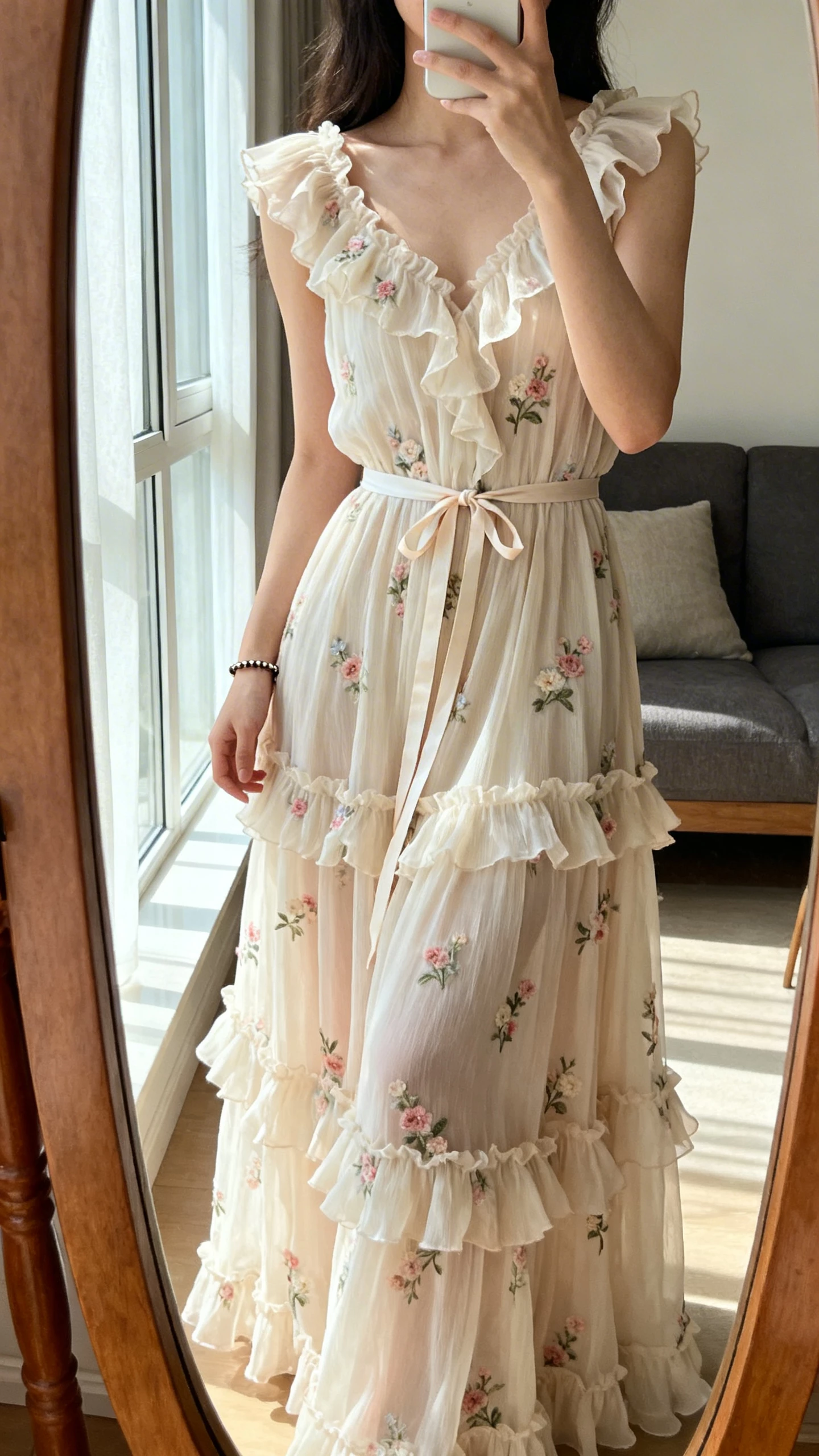 iPhone mirror selfie of a woman wearing a boho chiffon ruffle gown with barely-there floral appliqués, soft tiered ruffles, thin ribbon tie at waist, simple bracelet, face not visible, cropped at shoulders, living room mirror with afternoon window light, casual iPhone look