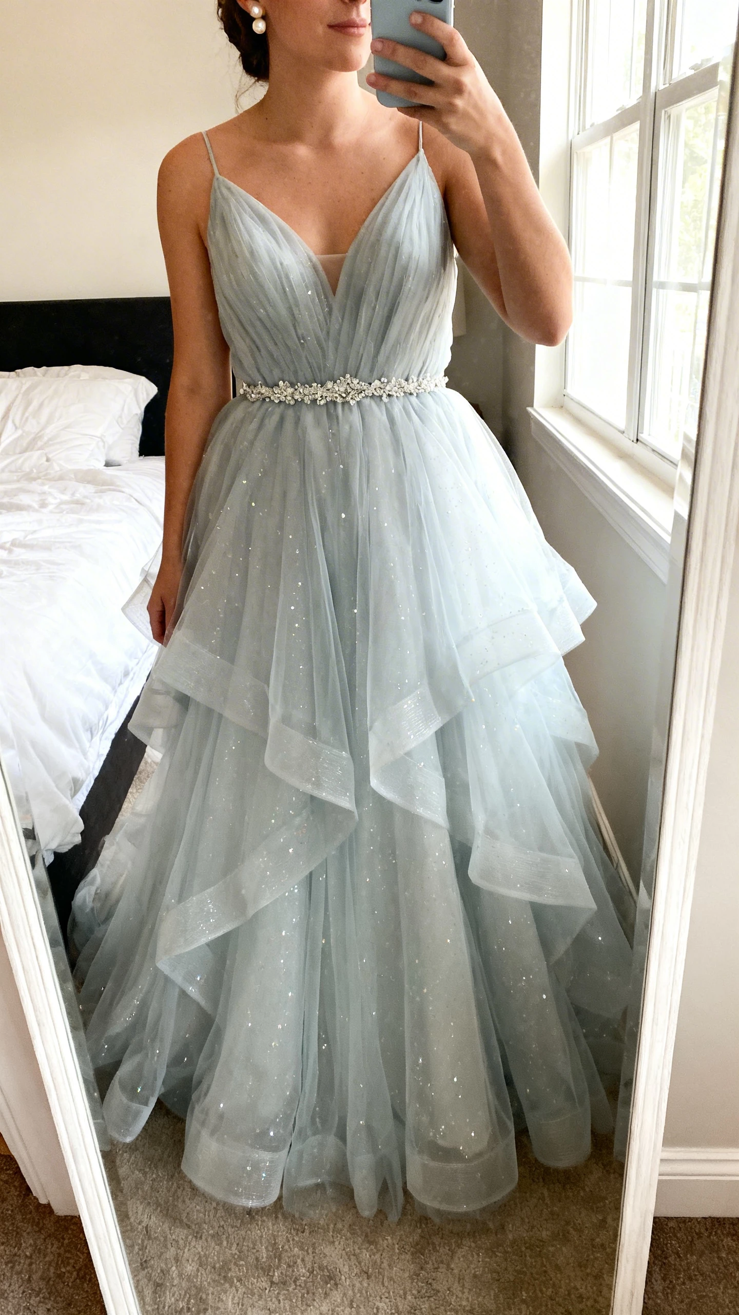 iPhone mirror selfie of a woman wearing a moonlit tulle princess wedding dress with layered airy tulle skirt, subtle shimmer/sparkle throughout, delicate beaded belt, simple pearl studs, face not visible, cropped at neck, bedroom full-length mirror, soft window light, natural iPhone photo quality