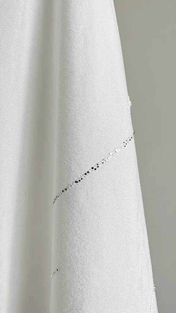 Close-up detail shot of white fabric with delicate embroidery and tiny silver sequins, fabric textures and stitching in sharp focus, neutral background, studio light, no text.