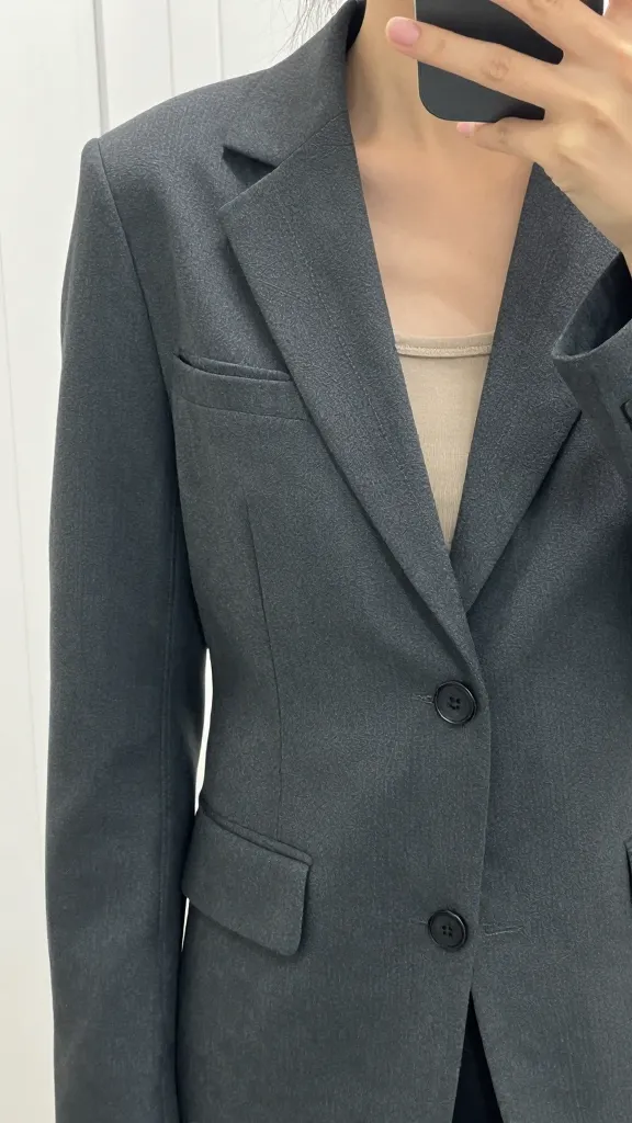 Close-up shot of a structured charcoal blazer worn as an outer layer, photographed in a mirror selfie from waist height to highlight lapels and stitching, against a clean white-walled room.