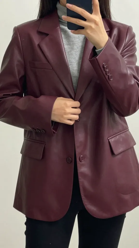 A close-up mirror selfie of a woman buttoning a deep burgundy leather jacket over a lightweight blazer, with the hem hitting at the waist and a hint of a gray turtleneck underneath, captured from chest to mid-thigh for a clean, structured look.