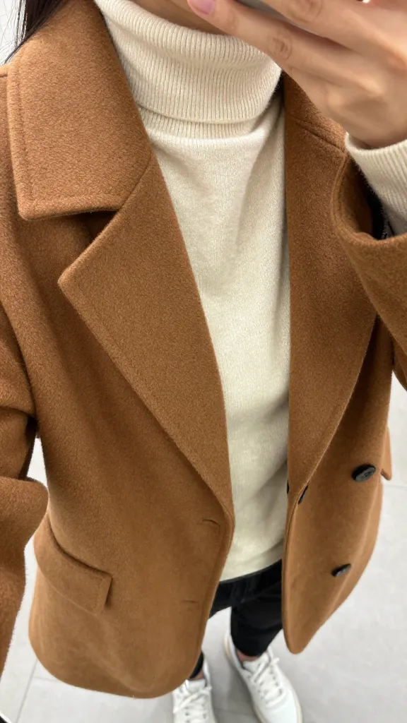 A close-up mirror selfie focusing on a brown caramel-colored jacket draped over shoulders, with a soft cream turtleneck and white sneakers in the frame, neutral blurred background, no faces visible.